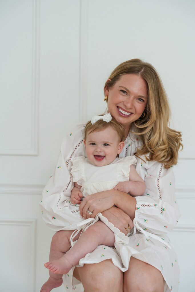 Michelle Schor in a white print dress holding daughter Zoe on her lap