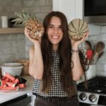 a woman with long dark hair standing in a kitchen holding a pineapple and melon on either side of her head
