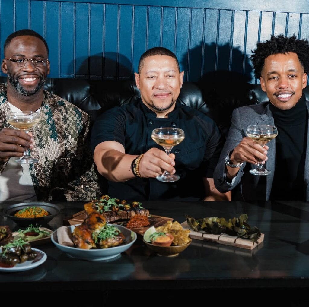 Three African-American men sit behind a table full of food at a restaurant raising cocktails in cheers