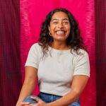 A young-adult Indian woman smiles directly at the camera sitting in front of a bright magenta fabric backdrop