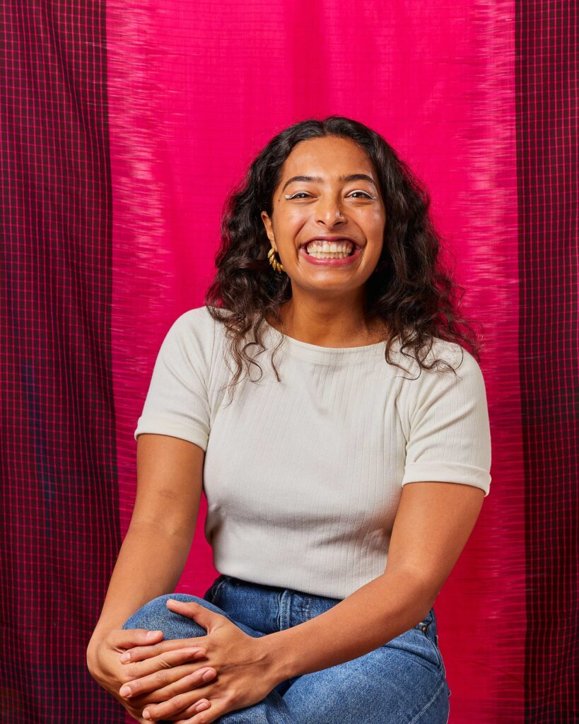 A young-adult Indian woman smiles directly at the camera sitting in front of a bright magenta fabric backdrop