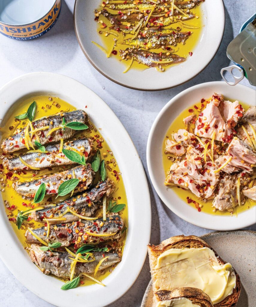 Plates of sardines and anchovies in olive oil with herbs and chili flakes, next to buttered toast