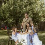A family of four sits around a decorated picnic table outdoors, all wearing white outfits