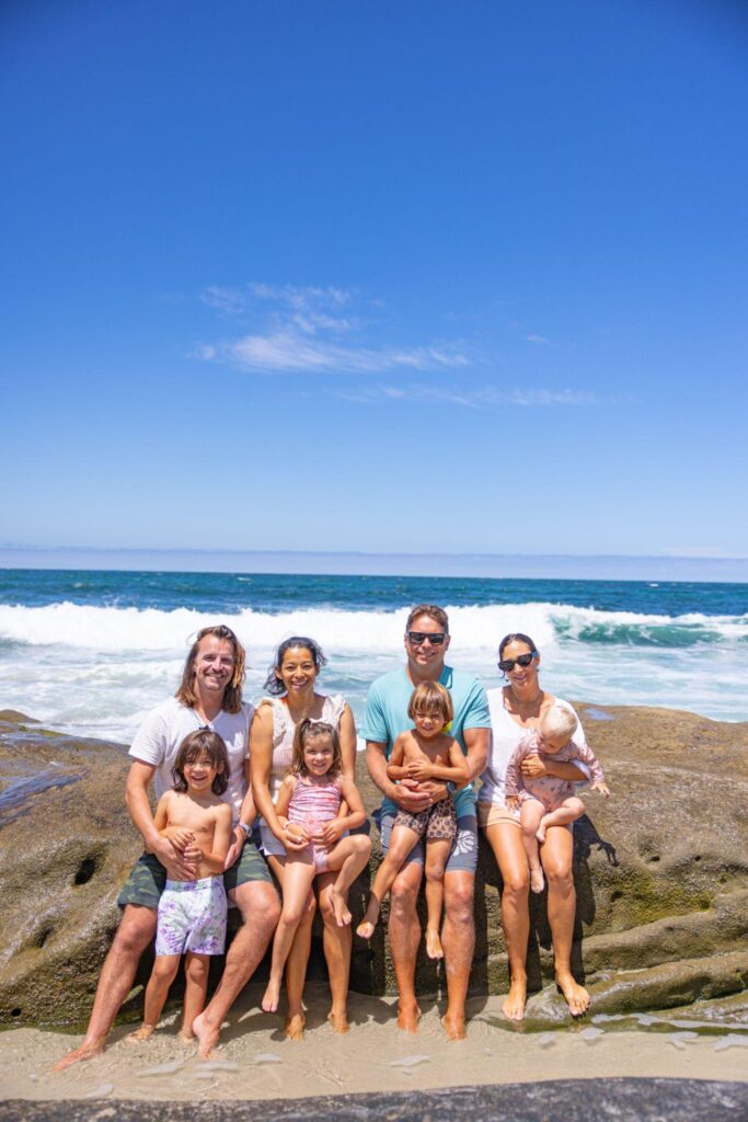 Torie Borrelli Hall, Eric Wolfinger and their families sitting together on rocks at the beach