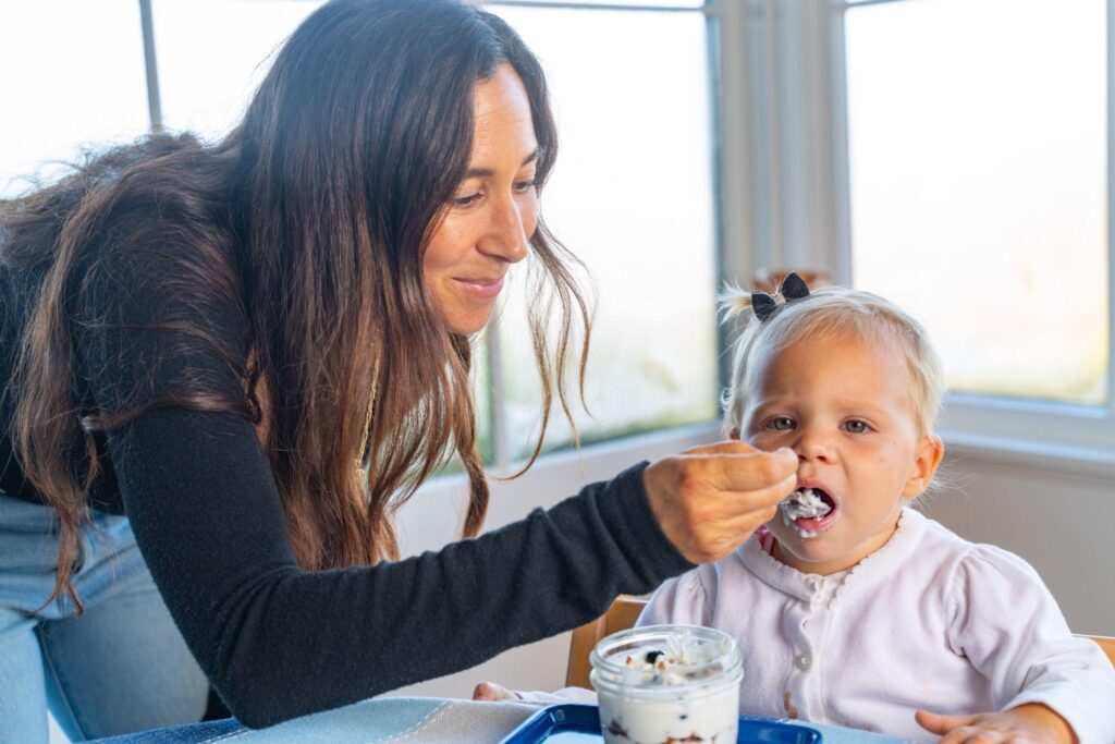Torie Borrelli Hall smiling as she spoon-feeds her baby daughter in a high chair