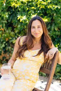 Torie Borrelli Hall, pregnant, in a yellow floral dress, sitting on a wooden bench in a garden and holding a glass of water