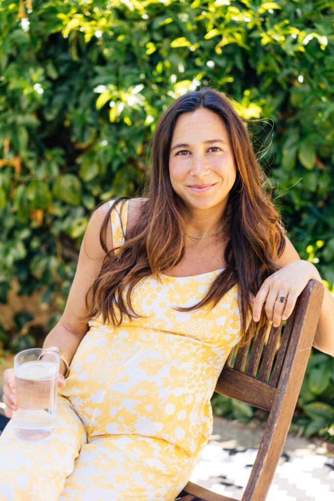 Torie Borrelli Hall, pregnant, in a yellow floral dress, sitting on a wooden bench in a garden and holding a glass of water